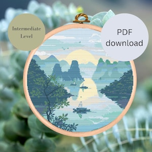 Pode incluir: Um padrão de ponto cruz de uma paisagem azul e verde com montanhas, um rio e um barco. O padrão está em um aro de bordado redondo e é rotulado como "Nível intermediário".