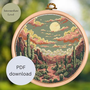 Könnte beinhalten: Kreuzstichmuster einer Wüstenlandschaft mit Vollmond, Kakteen und Bergen. Das Muster befindet sich in einem hölzernen Stickrahmen und ist mit "PDF-Download" und "Mittleres Niveau" beschriftet.