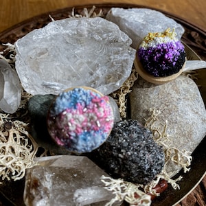 Könnte beinhalten: Zwei gestickte Anstecknadeln mit einer Vielzahl von Farben, darunter Pink, Blau, Weiß, Gelb, Schwarz und Lila. Die Nadeln liegen auf einem Bett aus Moos und klaren Quarzkristallen.