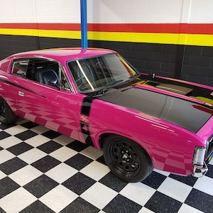 May include: A pink 1970s vintage muscle car with black racing stripes and black wheels. The car is parked in a garage with a black and white chequered floor.
