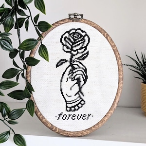 Puede incluir: Bastidor de bordado de punto de cruz ovalado con un diseño en blanco y negro de una mano sosteniendo una rosa. La palabra "forever" está cosida debajo de la mano. El bastidor tiene un marco de madera y un cierre metálico en la parte superior.