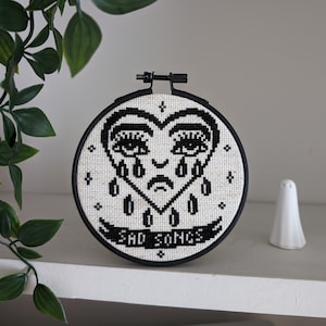 Puede incluir: Aro de bordado blanco y negro con un diseño de corazón llorando y el texto "Sad Songs".