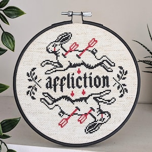 May include: A cross-stitch embroidery in a black hoop features two rabbits pierced by arrows, with the word "affliction" in the center. The design uses black and red thread on a cream-colored fabric, creating a gothic aesthetic.