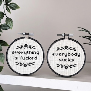 May include: Two black embroidery hoops with white fabric and black text that reads "everything is fucked" and "everybody sucks". The hoops are displayed on a white shelf with greenery in the background.