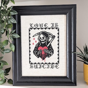 May include: Framed cross-stitch artwork in a black frame. The design shows a Grim Reaper figure holding a bleeding heart, with the text "LOVE IS SUICIDE" above and below. The colours used are black and red.