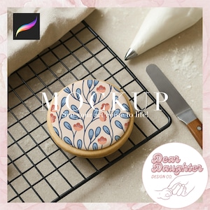 May include: A round cookie with a floral design featuring red flowers and blue leaves on a light beige background. The cookie is on a black wire rack, with a piping bag and spatula nearby. Text on the cookie says "MOCKUP" and "See your art come to life!"