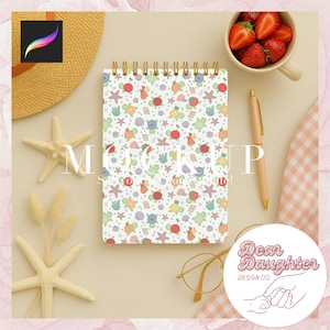 May include: A white spiral bound notebook with a colorful pattern of sea turtles, seashells, and starfish. The notebook is on a light brown surface with a straw hat, a bowl of strawberries, and a pen.