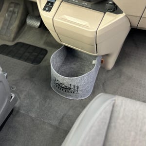 Puede incluir: Un cubo de basura para coche de fieltro gris con un diseño de montaña y árboles negros. El cubo es cilíndrico y cuelga debajo del salpicadero de un vehículo. El interior del coche es beige y gris.