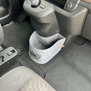 Peut inclure: Un organisateur de voiture en feutre gris avec un motif de montagne et d'arbres. L'organisateur est placé dans la console centrale d'une voiture, près du levier de vitesses et du tableau de bord. L'intérieur de la voiture est noir.