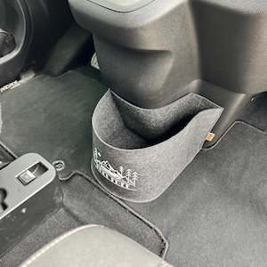 Può includere: Un organizer per auto in feltro grigio con un motivo di montagna e alberi. L'organizer è progettato per essere appeso in un'auto e ha una tasca per riporre oggetti. L'interno dell'auto è visibile sullo sfondo.