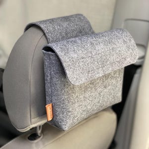 Può includere: Un organizer per auto in feltro grigio con chiusura a patta, attaccato a un poggiatesta per auto. L'organizer ha una forma rettangolare con una piccola etichetta arancione sul lato. Il sedile dell'auto è grigio.