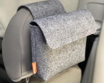 May include: A gray felt car organizer with a flap closure, attached to a car headrest. The organizer has a rectangular shape with a small orange tag that reads "AHLA". The car seat is gray.