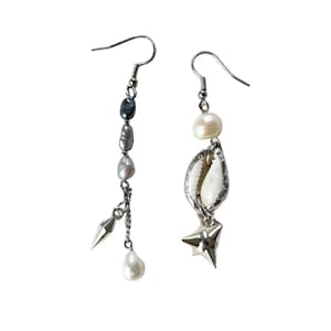 May include: A pair of mismatched drop earrings featuring silver-tone hooks. One earring has a vertical chain with dark grey and white pearls and a silver spike charm. The other earring features a white pearl, a white cowrie shell, and a silver star charm.