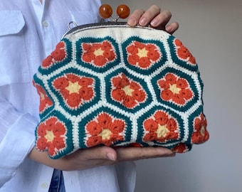 May include: A crocheted clutch with a floral pattern. The bag features a white background with orange and yellow flowers outlined in teal. The clasp has two brown, spherical accents.