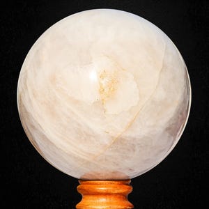 Può includere: Una grande sfera di cristallo sferica e traslucida con un aspetto marmorizzato beige chiaro e bianco. La sfera di cristallo poggia su una piccola base di legno tornito con una calda finitura marrone-rossastra. Lo sfondo è nero pieno.