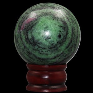 May include: A polished gemstone sphere, predominantly green with black and red veining, sits atop a dark wooden stand. The sphere's surface reflects light, showcasing its natural patterns. The stand is a rich, dark brown color.
