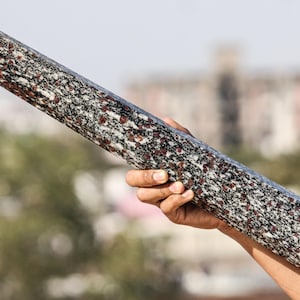 May include: A person holds a long, pointed, gray and red granite rod. The rod has a speckled pattern of red and black flecks.