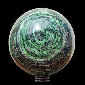 Könnte beinhalten: Eine polierte Kugel aus grünem, schwarzem und rotem Zoisit, die auf einem dunklen Holzsockel ruht. Die Oberfläche des Steins zeigt ein wirbelndes Farbmuster mit glänzendem Finish. Die Kugel ist ein dekorativer Gegenstand.