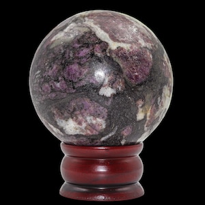 Può includere: Una sfera lucida con motivi viola, neri e bianchi, posta su un supporto in legno rosso scuro. La superficie della sfera ha un aspetto marmorizzato, con varie sfumature di viola e nero. Il supporto è di colore rosso scuro.