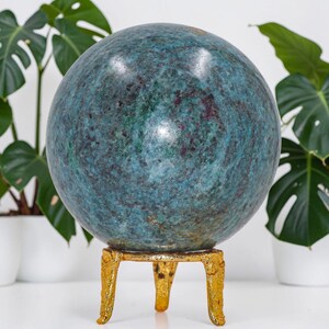 May include: A polished, spherical gemstone featuring a mottled texture with deep teal, blue, and dark red inclusions. The sphere rests securely on a three-legged, textured gold-coloured metal stand, providing a stable base for the round stone.