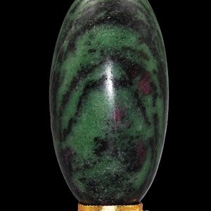 以下が含まれることがあります： 緑と黒の大理石模様の、磨かれた楕円形の石。小さな赤いインクルージョンがアクセントになっています。小さな金色の装飾スタンドに置かれ、黒い背景に配置されています。