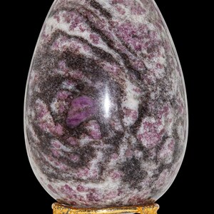Peut inclure: Un grand œuf en pierre polie, de forme ovale, avec un motif tourbillonnant de teintes blanches, gris foncé et rose-violet. L'œuf repose sur un petit support doré à trois pieds. Le fond est noir uni.