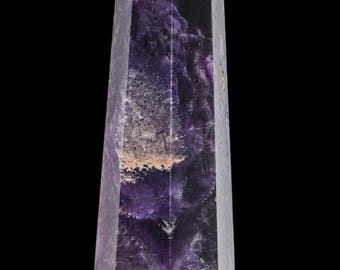 May include: A tall, faceted crystal obelisk with a pointed top. The obelisk features a blend of deep purple and clear sections, with a central area of tan and white inclusions. The crystal is set against a black background.