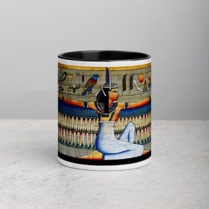 May include: White ceramic mug with a black interior and a detailed illustration of an Egyptian figure. The artwork features a seated woman with outstretched arms, adorned with hieroglyphs and vibrant colors, including blue, gold, and red.