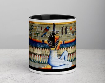 May include: White ceramic mug with a black interior and a detailed illustration of an Egyptian figure. The artwork features a seated woman with outstretched arms, adorned with hieroglyphs and vibrant colors, including blue, gold, and red.
