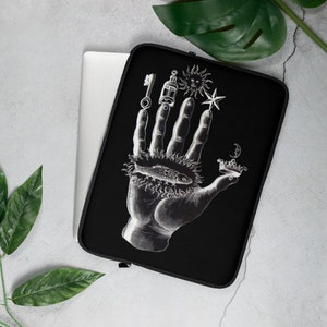 Puede incluir: Funda para port&aacute;til negra con una ilustraci&oacute;n blanca de una mano con varios s&iacute;mbolos, incluyendo una llave, una linterna, un sol, una estrella, un pez y una corona.