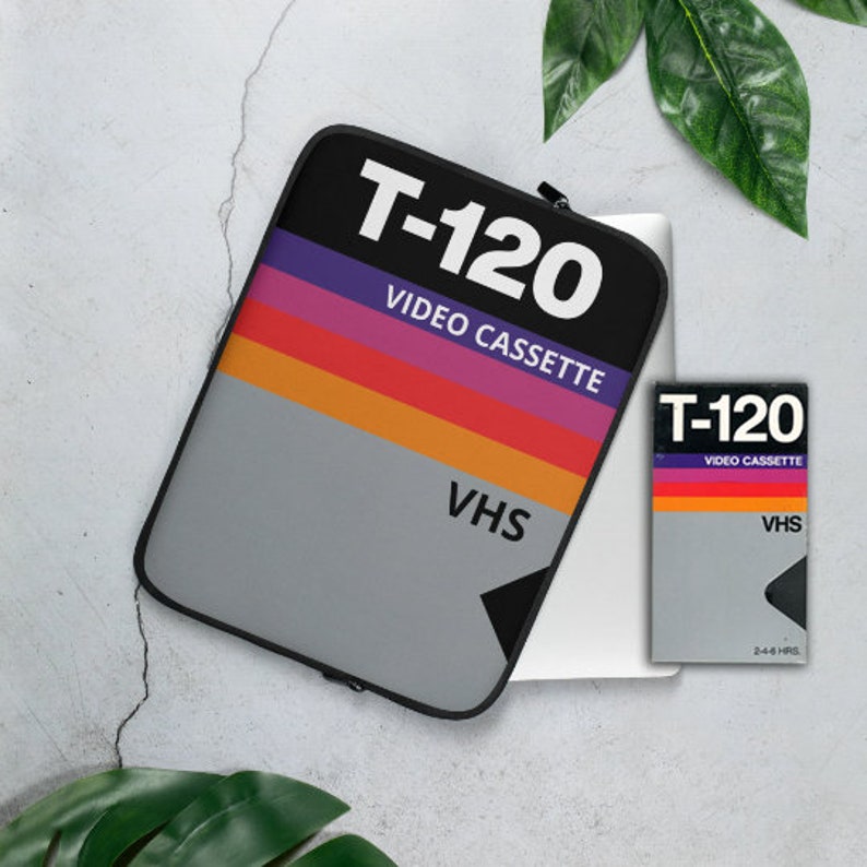 May include: A black laptop sleeve with a vintage VHS tape design. The sleeve features the text "T-120 VIDEO CASSETTE" and "VHS" in white. The sleeve is lying on top of a silver laptop.