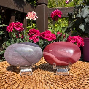 May include: Two smooth, oval-shaped stones displayed on clear plastic stands. One stone is a muted, dusty purple with subtle horizontal banding, while the other is a deep, solid red with a polished, reflective surface and faint white veins.