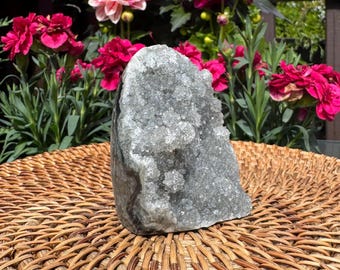 May include: A large, gray crystal geode stands on a woven wicker tray, with a backdrop of vibrant pink and red flowers. The crystal has a rough, textured surface and a natural, organic shape, perfect for home decor.