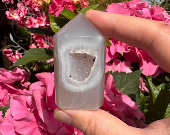 May include: A polished, semi-translucent light gray agate crystal tower with a hexagonal point. The stone features a central, hollowed-out cavity lined with small, sparkling white druzy crystals and subtle, concentric banding patterns.