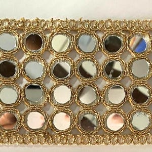 May include: Gold-colored trim with small, round, reflective mirror accents. The trim features a repeating pattern of mirrors set within a gold-colored, woven border. This decorative trim is suitable for various craft and sewing projects.