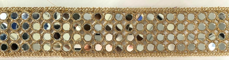 May include: Gold-colored trim with small, round, reflective mirror accents. The trim features a repeating pattern of mirrors set within a gold-colored, woven border. This decorative trim is suitable for various craft and sewing projects.