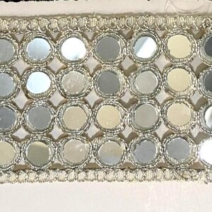 May include: A decorative trim featuring a repeating pattern of small, round, mirrored elements. The mirrored circles are set within a silver-colored, open-weave framework, and bordered by a delicate, silver-colored trim. The trim is set against a white background.