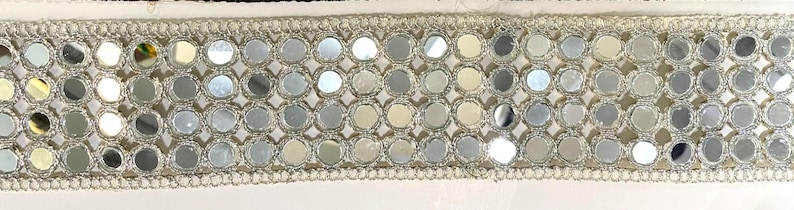 May include: A decorative trim featuring a repeating pattern of small, round, mirrored elements. The mirrored circles are set within a silver-colored, open-weave framework, and bordered by a delicate, silver-colored trim. The trim is set against a white background.