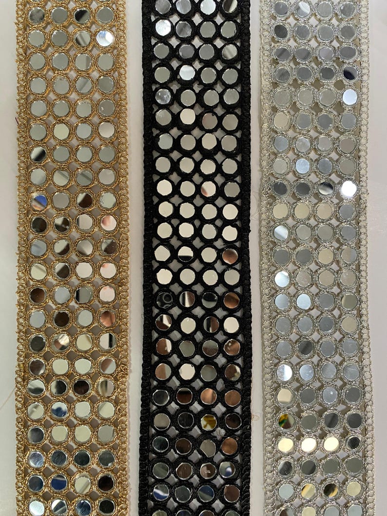 May include: Close-up of three decorative ribbons. The first is gold with small, round mirrors. The second is black with mirrors, and the third is silver with mirrors. All three ribbons have a textured, decorative edge.