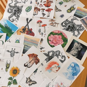 Peut inclure: Une collection d'estampes d'art présentant divers motifs, notamment une girafe, un tournesol, un cerf-volant, un champignon, une pieuvre, une guitare, une pivoine et un visage de femme. Les estampes sont sur du papier blanc avec de l'encre noire et sont disposées sur une surface en bois.