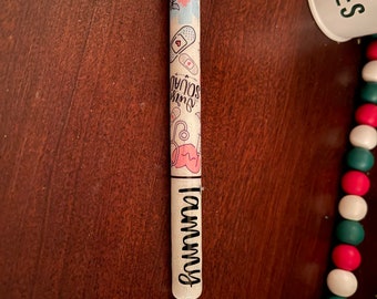 May include: A personalized pen with a black tip and a white barrel. The barrel is decorated with medical-themed illustrations, the words "Nurse Squad", and the name "Tammy". The pen is on a dark brown wooden surface.