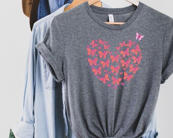 May include: A heather gray t-shirt with a pink and orange butterfly heart design. The shirt is hanging on a wooden hanger.