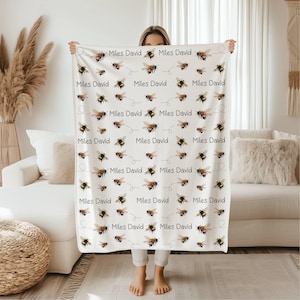 May include: Cream-coloured blanket with a repeating pattern of bees and the name "Miles David" in black. The bees are illustrated in black, brown, and yellow, with dotted flight paths. The blanket is held up by a person.