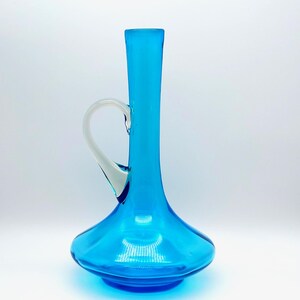 May include: A vibrant turquoise glass vase with a white handle. The vase has a bulbous base that tapers to a slender neck. The handle is a curved, clear glass. The vase is a decorative item.