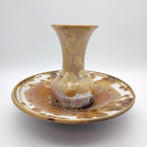 May include: A handcrafted ceramic vase and plate set. The vase is a light brown with a flared top and a bulbous base, featuring a speckled design. The plate is a matching brown with a wide rim, creating a cohesive decorative piece.
