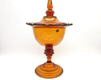 May include: Amber-colored glass compote with a lid. The compote features a stemmed base with a decorative pattern, a bulbous bowl, and a ruffled edge on the lid. The lid is topped with a pointed finial. The glass has a glossy finish.