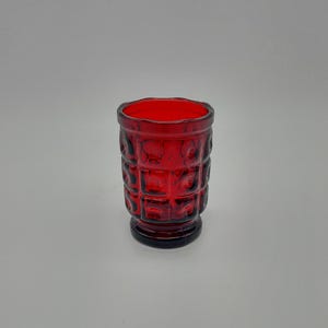 May include: A ruby red glass tumbler with a textured, square pattern. The vintage-style drinking glass has a thick base and a slightly flared rim. The glass is approximately 4 inches tall.