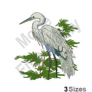 May include: White egret with a blue beak and legs standing in a green bush. The egret is facing to the right. The bush is in the background. The egret is a type of bird.