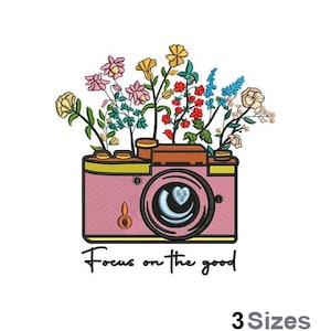 Op de afbeelding: Een roze camera met een bloemstuk erop. De camera heeft een lens met een halve maan en een hart erin. De tekst "Focus on the good" staat onder de camera.