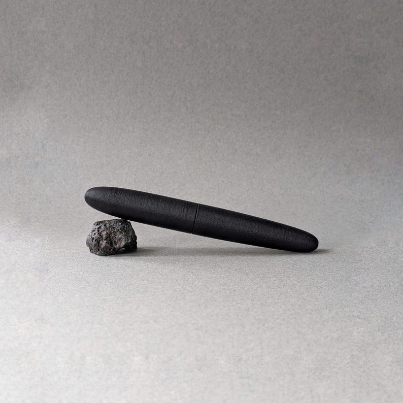 Peut inclure: Un stylo noir au fini mat repose sur une petite roche &agrave; la texture rugueuse. Le stylo a un design &eacute;l&eacute;gant et cylindrique avec une pointe arrondie. Le fond est gris neutre.